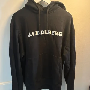 Svart hoodie från J.Lindeberg - Svart hoodie från J.Lindeberg med vit logga tryckt över bröstet. Klassisk modell med huva, snörning och stor magficka. Perfekt för en avslappnad och stilren look.