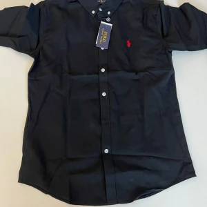 Svart skjorta från Ralph Lauren L - Klassisk svart skjorta från Ralph Lauren i bomull med vit knappstängning och röd broderad logga på bröstet. Skjortan har lång ärm och button down-krage, perfekt för en clean och stilren look.