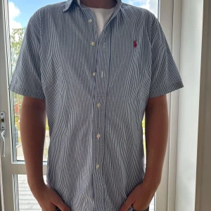 Randig skjorta från Ralph Lauren S - Klassisk kortärmad skjorta från Ralph Lauren i blåvitt randigt bomullstyg. Skjortan har knappar framtill, klassisk krage och röd broderad logga på bröstet. Passformen är classic fit och materialet känns svalt och skönt.
