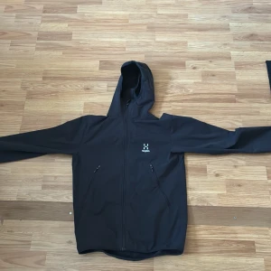Svart vindjacka från Haglöfs med huva - Svart vindjacka från Haglöfs med minimalistisk design, hel dragkedja och två snedställda fickor med dragkedja. Jackan har en justerbar huva och är perfekt för blåsiga dagar. Materialet är syntet och jackan har lång ärm den är ganska tunn. Priset är inte hugget i sten 