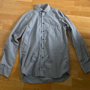 Rutig skjorta från Oscar - Snygg rutig skjorta från Oscar i svart och vitt, som ser grå ut på avstånd. Klassisk krage, långärmad med knappar vid ärmslut. 40 cm halsmått, Medium size. Skjortan är tillverkad i 100% bomull.