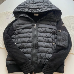 Svart Moncler jacka med huva - Snygg svart pufferjacka från Moncler med stickade ärmar och quiltad kropp. Jackan har huva, dragkedja framtill och en praktisk ficka med dragkedja på ärmen. Perfekt för kyliga dagar och har klassisk Moncler-logga på ärmen.