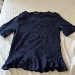 Mörkblå blus med volang från Gina Tricot - Supersöt mörkblå blus från Gina Tricot med smockad framsida och baksida. Toppen har korta ärmar och en volangkant längst ner som ger en extra touch. Perfekt för dig som gillar en lite sötare stil och vill ha något som sticker ut.