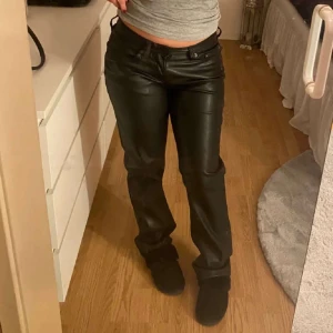 Svarta raka byxor i skinnimitation - Säljer ett par svarta byxor i skinnimitation med rak passform och klassisk femficksdesign. Byxorna är lowwaist och stängs med dragkedja och knapp. Perfekta skinbyxorna, använt en gång. 