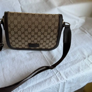 Gucci brun axelväska med GG-mönster - Säljer en snygg axelväska från Gucci i beige och brunt med klassiskt GG-mönster. Väskan har justerbar axelrem i textil, detaljer i brunt skinn och innerfack med dragkedja. Perfekt för dig som gillar stilrena och ikoniska accessoarer.