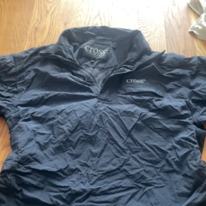Svart vindjacka från Cross Sportswear - Svart vindjacka från Cross Sportswear i storlek S. Jackan har halv dragkedja framtill, meshfoder och justerbara band i midjan. Perfekt för blåsiga dagar och enkel att slänga på sig när du är på språng.