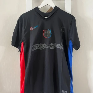 FC Barcelona x Cactus Jack Nike tröja - Snygg svart FC Barcelona x Cactus Jack fotbollströja från Nike. Tröjan har korta ärmar, röda och blå paneler på sidorna, klubbmärke på bröstet och tryck med Cactus Jack-logga. Materialet är lätt och andas, perfekt för match eller träning.