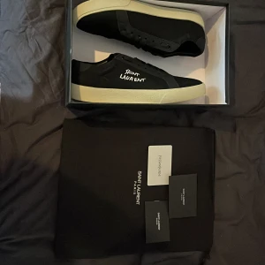 Svarta Saint Laurent sneakers - Snygga svarta sneakers från Saint Laurent med vit sula och handskriven logga på sidan. Skorna har klassisk snörning och rund tå. Kommer med originalkartong, dustbag och äkthetskort. Perfekta för dig som gillar stilrena och exklusiva sneakers.
