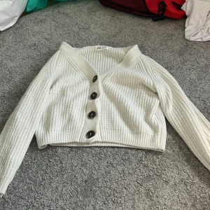 Vit stickad kofta från H&M - Supermysig vit stickad kofta från H&M med stora svarta knappar framtill. Koftan har v-ringning, ribbade muddar och är croppad i modellen. Perfekt att slänga över en t-shirt eller linne när det är lite kyligt ute. En pytte liten svart fläck på armen, syns knappt.