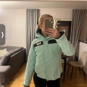 Ljusblå vindjacka från Helly Hansen - Snygg ljusblå vindjacka från Helly Hansen med dragkedja och hög krage. Jackan har svarta detaljer och logotyp på bröstet och ärmen. Perfekt för blåsiga dagar. Använd en gång