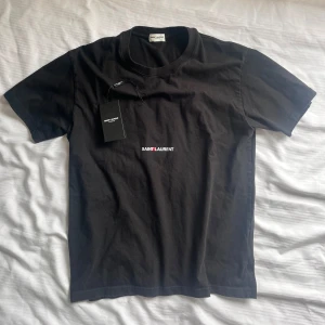 Svart Saint Laurent t-shirt - Svart t-shirt från Saint Laurent med klassisk rund hals och korta ärmar. Diskret logga i vitt och rött framtill. Tillverkad i mjuk bomull för en skön känsla och snygg passform. Perfekt för dig som gillar stilrena designer och exklusiva märken.