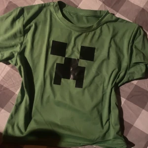 Grön Minecraft Creeper t-shirt - Säljer en grön t-shirt med det ikoniska Creeper-ansiktet från Minecraft tryckt i svart på bröstet. T-shirten är i mjuk bomulls- och polyestermix och har klassisk rund hals och korta ärmar. Perfekt för dig som älskar gaming och vill sticka ut med en cool look. Det står inte vilken storlek det är men skulle gissa att det passar runt alla (beror på om man vill ha baggy tröja eller crop-top)