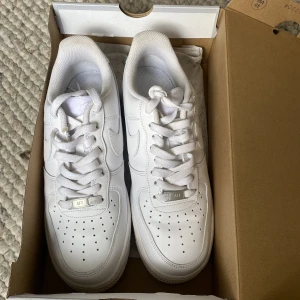 Nike Air Force 1 vita sneakers - Klassiska Nike Air Force 1 sneakers i helvitt skinn med snygga detaljer och perforerad tå. Lågt skaft, rund tå och platt sula. Perfekta för dig som gillar en clean och tidlös look. Skorna har snörning och ikonisk Nike Swoosh på sidorna.