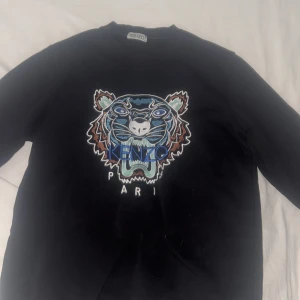 Svart Kenzo tröja med tigerbrodyr - Svart tröja från Kenzo med ikonisk broderad tiger i blått, vitt och brunt framtill. Klassisk rund hals och långa ärmar. Perfekt statement piece för dig som vill sticka ut med en snygg logga och cool design. Orginal priser är 1100!❤️ inga skador,