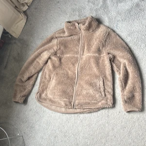 Beige teddyjacka från 157, storlek M - Mysig beige teddyjacka från 157 i storlek M. Jackan har hel dragkedja framtill, hög krage och två sidofickor. Perfekt för kyliga dagar när du vill vara både varm och snygg. Den fluffiga ytan ger en trendig look som passar till det mesta.