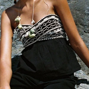 Svart bandeau tubtopp med broderi - Supersnygg svart bandeau tubtopp med vit broderad detalj upptill. Toppen är axelbandslös och har en lätt utsvängd passform nedtill. Perfekt för varma dagar när du vill sticka ut lite extra.