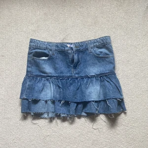 Blå jeanskjol med volanger - Säljer en blå jeanskjol med volangdetaljer och råa kanter. Kjolen har klassiska fickor fram och bak, bälteshällor och dragkedja. Perfekt för dig som gillar en avslappnad och trendig stil. Helt ny !