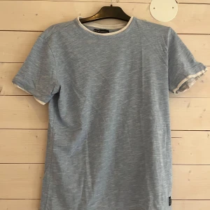 Ljusblå t-shirt från S.C.W. - Ljusblå t-shirt från S.C.W. med vita detaljer vid halsringning och ärmslut. T-shirten har en avslappnad passform och är tillverkad i mjuk bomull. Perfekt för dig som gillar en clean och enkel stil.