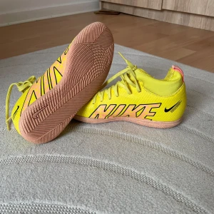 Gula Nike fotbollsskor - Snygga Nike  fotbollsskor i neon-gult med stora svarta och rosa loggor på . Skorna har snörning, platt ljusbrun sula och är gjorda i syntetmaterial. Perfekta för inomhusspel och sticker verkligen ut på planen. Storleken 36,5