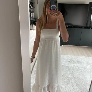 Vit långklänning med volangkant - Superfin vit långklänning med smala axelband och volangkant nertill. Klänningen har en söt rosettdetalj i ryggen och är lätt strukturerad i tyget. Endast använd en gång. Hör av dig om du har några frågor eller funderingar!🩷