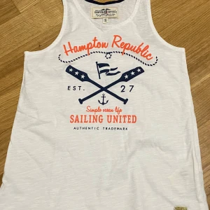 Vit linnetopp från Kappahl  - Snygg vit linnetopp från Kappahl med marinblå och orange tryck framtill. Toppen har ett sportigt seglingsinspirerat motiv med korsade åror, flagga och ankare. Rund halsringning med marinblå kant. Perfekt för en avslappnad och cool stil.