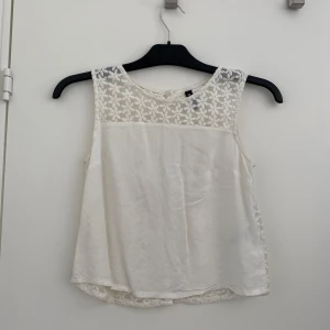 Vit blommig ärmlös topp H&M. Stl 34 - Superfin vit ärmlös topp från H&M Divided med broderade blommor i spets över axlar och rygg. Knappar längs ryggen och lätt A-linjeformad passform. Perfekt till jeans eller kjol när du vill ha en söt och somrig look. Liten fläck på nedre delen.     Hör av dig vid frågor!                                           Köparen står för frakten