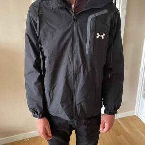 Svart vindjacka från Under Armour - Svart vindjacka från Under Armour med huva och dragkedja framtill. Jackan har en sportig look med logga på bröstet och diskreta detaljer. Tillverkad i lätt polyester med meshfoder för extra komfort. Perfekt för blåsiga dagar och enkel att matcha med allt.