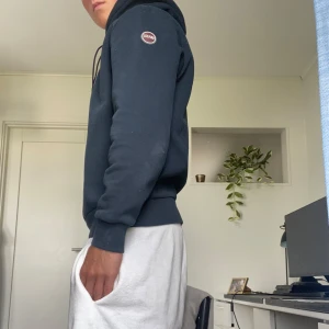 Mörkblå hoodie från Colmar - Snygg mörkblå hoodie från Colmar med klassisk huva och dragsko. Tröjan har en stilren look med ribbade muddar och Colmar-logga på ärmen. Perfekt för dig som gillar en enkel men trendig stil.