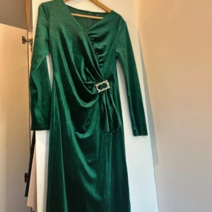 Grön sammetsklänning från SHEIN - Mörkgrön långklänning i sammet från SHEIN med omlott-design, lång ärm och snygg v-ringning. Klänningen har en dekorativ pärlspänne i midjan och dragkedja bak. Perfekt för dig som vill sticka ut med en elegant look.
