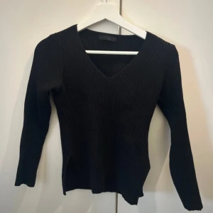 Svart ribbad stickad v-ringad tröja - Säljer en svart stickad tröja med v-ringning och lång ärm. Tröjan är ribbad och har en figurnära passform som ger en clean och stilren look. Perfekt att matcha med jeans eller kjol för en enkel men snygg outfit.