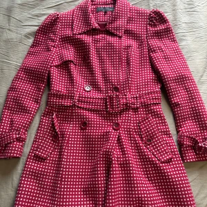 Prickig röd  trenchcoat från Next - Snygg röd trenchcoat från Next med vita prickar, klassisk krage och bälte i midjan. Jackan har långa ärmar, knappar framtill och coola detaljer vid ärmslut och fickor. Perfekt statement-plagg för dig som vill sticka ut med färg och mönster.