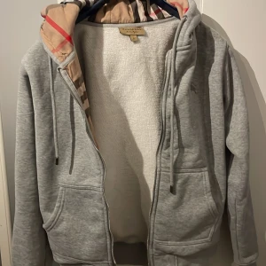 Grå hoodie från Burberry - Grå hoodiejacka från Burberry med dragkedja, fickor och klassiskt rutigt foder i huvan. Hoodien har snörning i huvan och är fodrad med mjuk fleece på insidan. Perfekt för dig som vill ha en stilren och bekväm tröja med lyxig känsla.