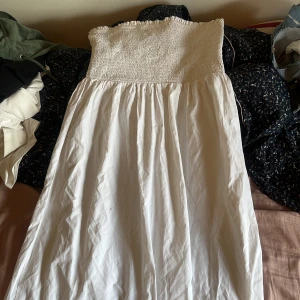 Vit långklänning från BikBok - Supersöt vit långklänning från BikBok med smockad överdel och volangkant upptill. Klänningen är axelbandslös och har en luftig, flowy kjol som passar perfekt till varma dagar. Enkel och stilren design som är lätt att matcha.