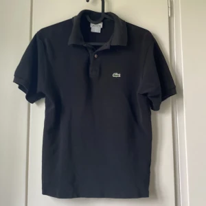 Svart piké från Lacoste - Klassisk svart pikétröja från Lacoste med korta ärmar och den ikoniska gröna krokodilloggan broderad på bröstet. Tröjan har krage och knappar framtill, tillverkad i mjuk bomull som andas. Ca 12 till 14 år.          Originalpris : 750 - 900 kr