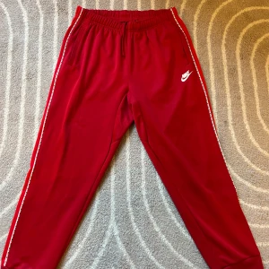 Röda Nike mjukisbyxor med logga - Snygga röda mjukisbyxor från Nike med vit logga på låret och vita detaljer längs sidorna. Byxorna har resår och snörning i midjan, ribbade muddar vid bensluten och är gjorda i 100% polyester. Perfekta för chill eller träning.