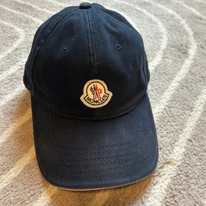 Mörkblå keps från Moncler - Snygg mörkblå keps från Moncler med klassisk böjd skärm och broderad logotyp framtill. Kepsen är tillverkad i bomull och har ventilerande hål upptill. Insidan är fodrad med rött tyg för en extra detalj. Perfekt för dig som gillar stilrena accessoarer.