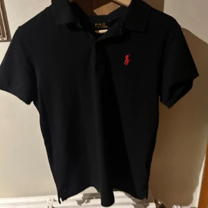 Svart pikétröja från Polo Ralph Lauren - Klassisk svart pikétröja från Polo Ralph Lauren med röd broderad logga på bröstet. Tröjan har korta ärmar, krage och knäppning framtill. Tillverkad i mjuk bomull med lite stretch för skön passform. Perfekt till jeans eller shorts.