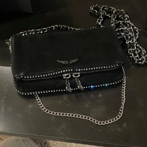 Svart axelväska från Zadig & Voltaire - Snygg svart axelväska i mocka från Zadig & Voltaire och snygga detaljer med stenar. Storlek ”nano”. Den är väl använd. Defekter förekommer som sminkfläckar men går att få bort säkert, två stenar har lossnat men inget som syns alls. Dustbag medföljer inte eftersom hon hade tappat bort den när jag köpte här! 