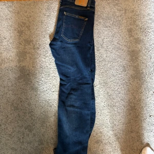 Mörkblå jeans från Nudie Jeans, 29/30 - Snygga mörkblå jeans från Nudie Jeans med klassiska kontrastsömmar och bakficka med logga. Modellen har normal passform och är tillverkad i slitstarkt bomullsjeans. Perfekta för dig som gillar stilrena och tidlösa jeans. Nypris 1600
