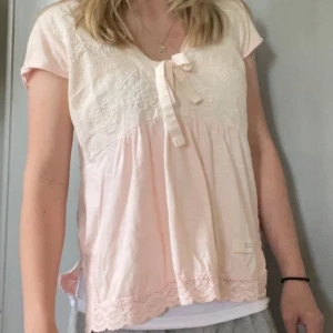Ljusrosa blus från Odd Molly - Supersöt ljusrosa blus från Odd Molly med broderade detaljer upptill, knytning vid bysten och spetskant nertill. Blusen har korta ärmar. Storlek 1. 