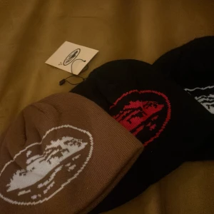 Stickade mössor med grafiskt tryck - Tre stickade mössor i brunt, svart och svart med grafiskt motiv framtill. En mössa har vitt tryck, en har rött och en har vitt motiv. Perfekt accessoar för att hålla värmen och addera stil till din outfit.