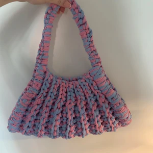 Virkad axelväska i rosa och blått - Superfin handgjord axelväska med chunky virkat mönster i pastellrosa och ljusblått. Väskan har en bred axelrem och är gjord i mjukt material som ger en cool och unik look. Perfekt för dig som vill sticka ut med accessoarer!