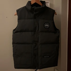 Grå väst från Canada Goose (A-Kop) - Säljer en grå väst från Canada Goose med hög krage och två stora fickor framtill. Västen har broderad logga på bröstet och stängs med knappar. Aldrig använd!!