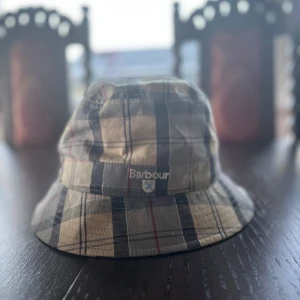 Barbour fiskehatt - Snygg bucket hat från Barbour i klassiskt rutig design med beige, svart och röda toner. Hatten är tillverkad i 100% bomull och har broderad Barbour-logga framtill. Perfekt accessoar för att lyfta din stil under soliga dagar. Säljer pga för den är för liten. Helt ny. 