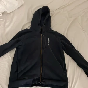 Svart Sail Racing vindjacka med huva - Svart vindjacka från Sail Racing med huva och hel dragkedja. Jackan har en bröstficka med dragkedja och Sail Racing-logga på bröstet. Tillverkad i mjukt syntetmaterial, perfekt för blåsiga dagar. Snygg och sportig look med långärmad passform.