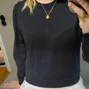 Svart sweatshirt med broderade ärmar - Svart sweatshirt med rund hals och långa ärmar. Ärmarna har broderade detaljer i form av små blommor och lätt puff vid axlarna. Tröjan har ribbade muddar vid ärmslut och nederkant. Perfekt för dig som gillar en enkel men ändå unik look.