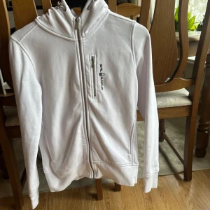Vit hoodiejacka från Sail Racing XS - Vit hoodiejacka från Sail Racing med dragkedja framtill och på bröstfickan. Jackan har huva med snörning och broderad logga på bröstet. Mjuk bomullskänsla och långärmad modell, perfekt för chill dagar eller när du vill ha en clean look.