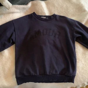 Mörkblå sweatshirt Amour Gina Tricot - Mörkblå sweatshirt från Gina Tricot med texten 'AMOUR' i svarta pärlor framtill. Klassisk rund halsringning, ribbade muddar vid ärmslut och nederkant. Perfekt för dig som gillar enkel men snygg stil.