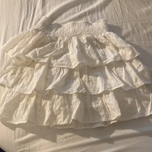 Vit volangkjol från Lindex - Supersöt vit kortkjol från Lindex med flera lager volanger och smock i midjan. 