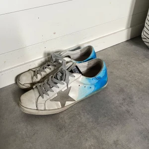 Golden Goose Superstar - Golden Goose Superstar sneakers i vitt skinn med grå stjärndetalj på sidan och handmålat blått parti baktill. Grå snörning med unika detaljer och text på hälen. Perfekta för dig som gillar statement-sneakers. Skriv om du har frågor 🤩